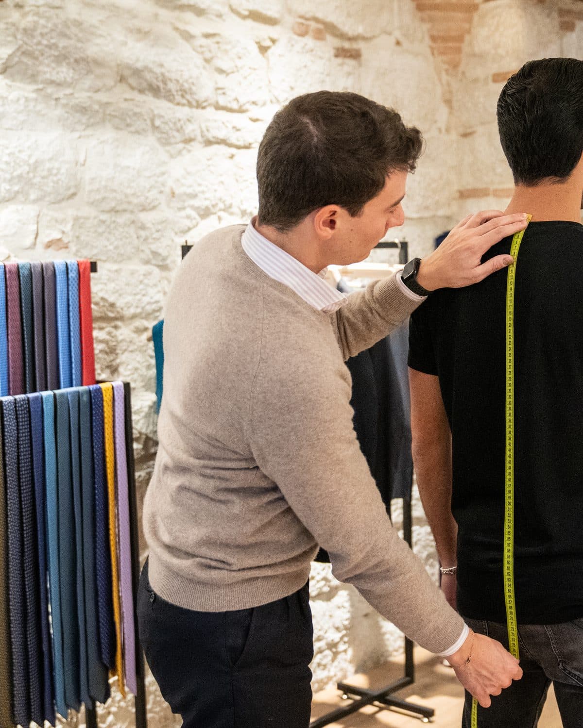A tailor measuring a customer beside a rail of Italian cloth swatches during an in-store fitting appointment